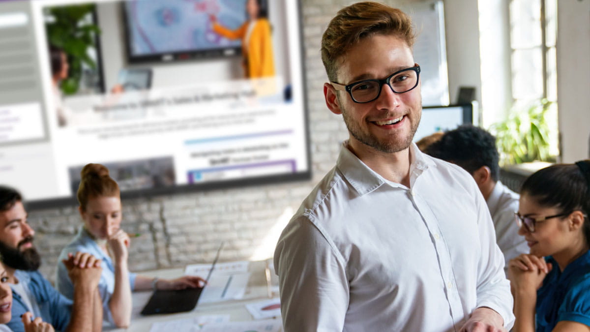 Professional standing confidently in front of colleagues during a meeting, with a SMART Board in the background being used for a presentation.